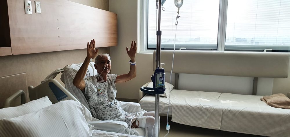The keeper's brother-in-law in a hospital bed in 2022, frail but with both hands raised in praise