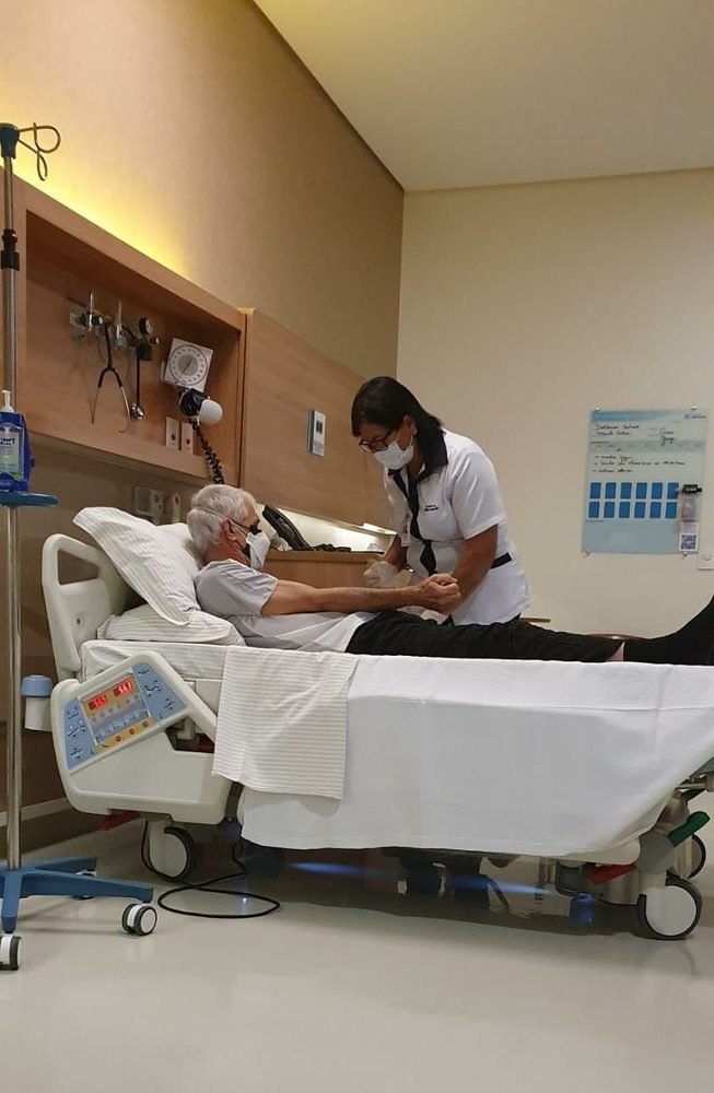 The brother-in-law in 2025, resting in his hospital bed while a nurse tends to him