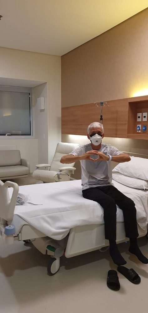 The brother-in-law in 2025, sitting up in his hospital bed making a heart shape with his hands